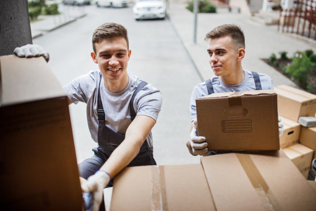 Two young handsome smiling movers wearing uniforms are unloading the van full of boxes. House move, mover service